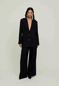 Kobieta w oversize'owej czarnej marynarce i szerokich spodniach z długimi białymi koralikowymi kolczykami na tle jasnym.