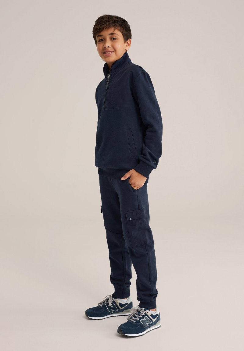 Temno modra flis majica s polovičnim zapenjanjem in stranskimi žepi, v kombinaciji z usklajenimi hlačami za tek, ki imajo cargo žepke in gladko teksturo.