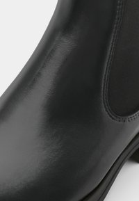 Svart läderankelboot med en polerad yta, elastiska sidopaneler och en mjuk textur. Har en rund tå och låg klack.