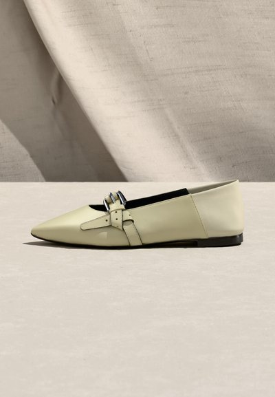 Chaussure plate en cuir couleur crème avec un bout pointu et des brides croisées à boucle, vue de profil sur un fond en tissu neutre.
