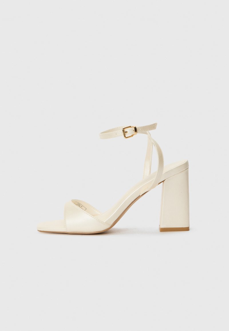 Cream-colored heeled sandals with an open toe, thick block heel, thin ankle strap, and subtle sheen finish. Buckle accent on strap.