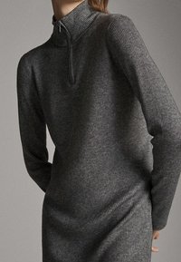 Szara dzianinowa sukienka z prążkami, z wysokim kołnierzem i pół zamkiem błyskawicznym. Materiał wydaje się być fakturowany i blisko ciała, z długimi rękawami.