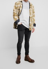 Bunda s maskovacím vzorem v béžové a olivové barvě, šedý pletený svetr, černé roztrhané úzké džíny, hnědé kožené Chelsea boty.