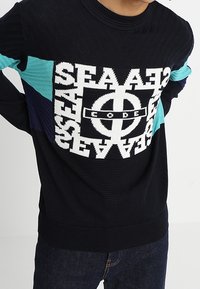 Gestickter Pullover in Marineblau mit einem auffälligen weißen und türkisfarbenen grafischen Design auf der Brust. Rippstruktur und einfarbige Ärmel.