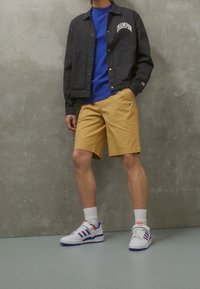 Szara kurtka z logo, niebieska koszula, żółte szorty i białe trampki z niebieskimi i czerwonymi akcentami. Gładki materiał, casualowy krój.