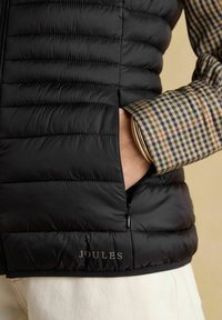 Fekete puffermellény steppelt textúrával, oldalzsebbel és Joules márkázással. Alatta kék és barna árnyalatokban mintás ing látható.