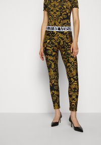 Schwarz-goldene gemusterte Leggings mit enger Passform und einem weißen Gummibund. Kombiniert mit schwarzen High Heels.