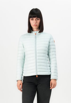 Vrouw met steil donker haar, gekleed in een lichtblauwe gewatteerde jas met oranje ritslipjes en zwarte broek, staand tegen een witte achtergrond.
