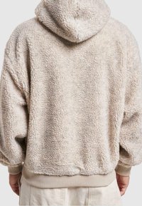 Felpa con cappuccio in pile beige e testurizzato, con polsini e vita a costine, vista da dietro su una persona che indossa pantaloni chiari.