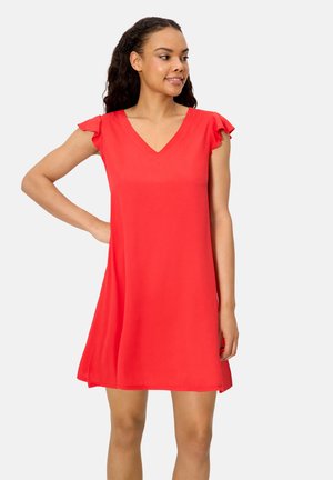 Mujer con un vestido rojo sin mangas con hombros con volantes, de pie con una mano en la cadera, sonriendo y mirando hacia un lado.