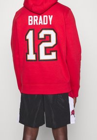 Roter Hoodie mit "BRADY" und "12" in Weiß und Orange auf dem Rücken. Mit Kängurutasche und verstellbarem Kordelzug an der Kapuze. Schwarze Shorts.