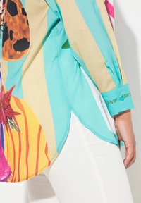 Blouse multicolore avec des motifs abstraits audacieux en turquoise, beige et jaune. Tissu lisse avec un ourlet arrondi et des détails de poignet.