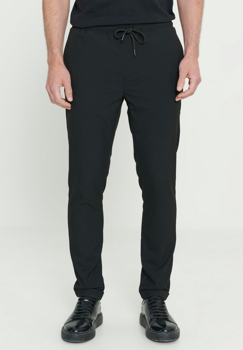 Zwarte taps toelopende broek van een gladde stof met een elastische tailleband en trekkoord. Bevat zijzakken en aansluitende boorden.