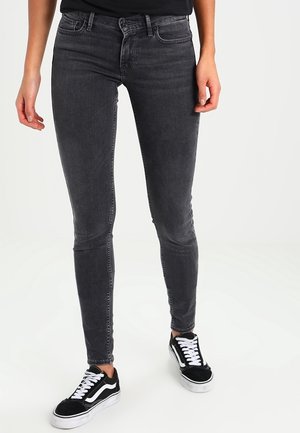 Person wearing black skinny jeans and black sneakers with white laces and soles, standing on a white background.