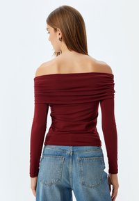 Bordeaux off-the-shoulder top med samlet detalje, lange ærmer og en tætsiddende pasform, parret med klassiske blå denimjeans.