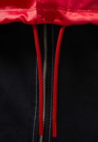 Lähikuva punaisen hupun nyöreistä ja metallisesta vetoketjusta mustassa kankaassa, jonka yläreunassa on punainen painikenappi.