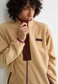 Beige fleece jacket featuring a high collar, brown zip, and purple accents. Logo on the left chest and elastic cuffs. Two side pockets.