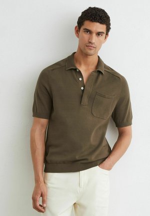 Man draagt een olijfgroen poloshirt met korte mouwen, knopen en een borstzak, gecombineerd met lichte broek.
