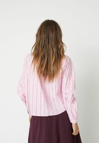 Blusa a righe rosa con maniche lunghe e taglio cropped. Tessuto testurizzato; sottili righe verticali aggiungono dettaglio. Indossata con una gonna scura.
