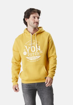 Homme portant un sweat à capuche jaune avec des textes et graphiques blancs de style vintage, debout les mains détendues le long du corps, regardant vers la droite.