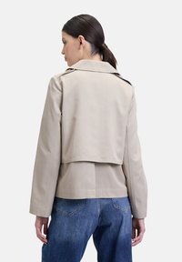 Beige jas met een cropped ontwerp, voorzien van een grote kraag, achterste split en lange mouwen. Dragen over blauwe denim jeans.