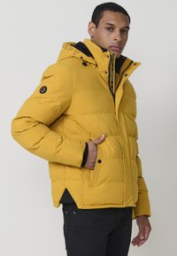 Giacca a vento gialla con colletto alto, dettagli neri e zip frontale. Presenta una patch con il logo sulla manica e tasche laterali.
