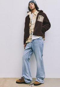 Brązowa kurtka polarowa z zamkiem, żółta koszula w kratę, jasnoniebieskie dżinsy i brązowe buty ze zdobnymi sznurówkami. Prosty, casualowy strój.