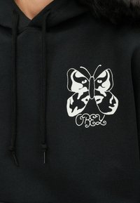 Fekete kapucnis pulóver fehér pillangó grafikával és "OBEY" felirattal, húzózsinórral ellátott kapucnival és puha textúrával.