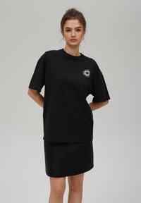 Crna oversized majica od pamuka s okruglim grafičkim uzorkom sunca na prsima. Uparen s crnom kožnom mini suknjom. Glatka tekstura.