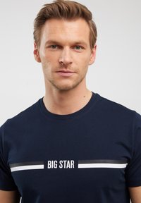 Tamsiai mėlyna medvilninė marškinėliai su trumpomis rankovėmis, apvalia iškirpte ir grafiniu spaudiniu, kuriame baltais ir pilkais dryžiais užrašyta "BIG STAR" per krūtinę.