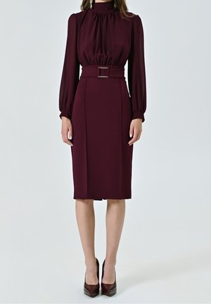 Mujer con un vestido midi burdeos de manga larga y cinturón, con cuello alto, combinado con tacones a juego de punta fina y pendientes colgantes.