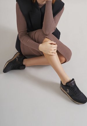 Personne assise en tailleur portant une robe brune côtelée, un gilet matelassé noir et des baskets noires avec des semelles beiges sur un sol clair.