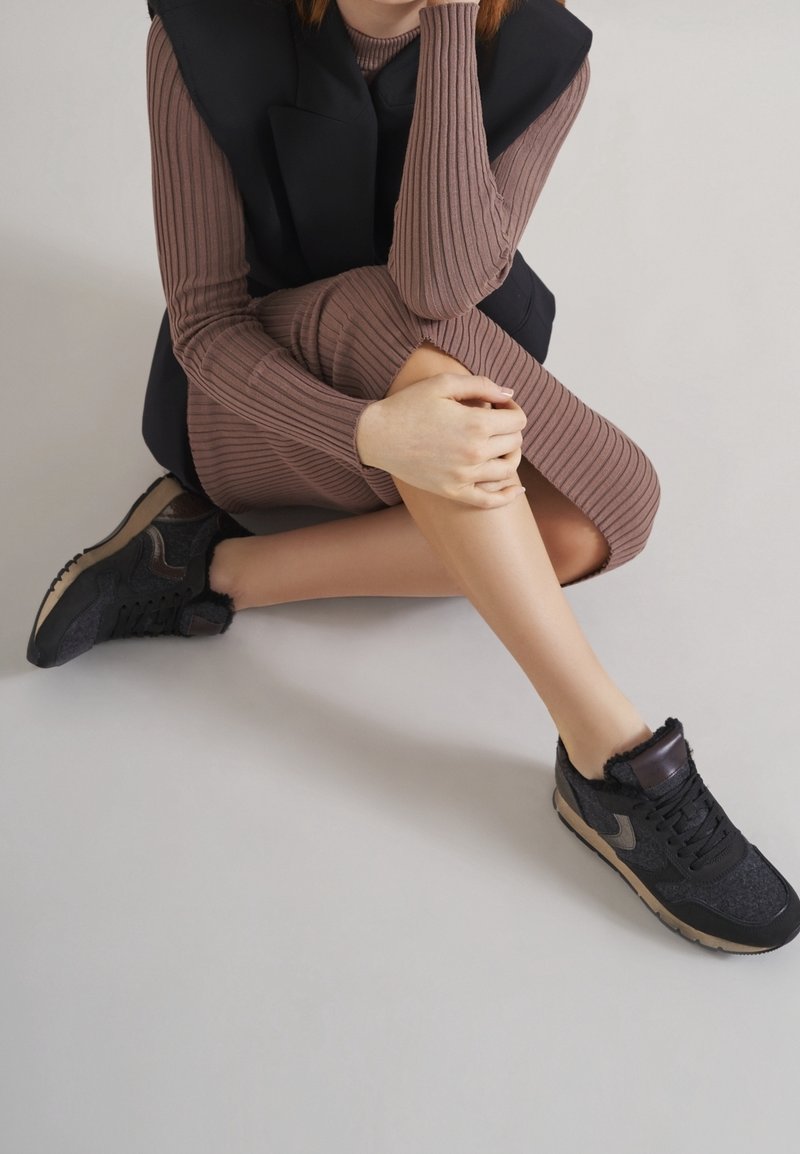 Personne assise en tailleur portant une robe brune côtelée, un gilet matelassé noir et des baskets noires avec des semelles beiges sur un sol clair.