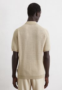 Beige short-sleeve polo shirt featuring a textured knit design, ribbed collar, and cuffs, with a straight hem and label at the side seam.