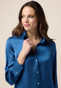 Blusa blu in satin con maniche lunghe, colletto a punta e lucentezza delicata. Presenta bottoni dorati lungo la parte anteriore e dettagli ai polsini.