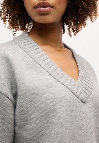 Κοντινό πλάνο ενός ατόμου που φοράει ανοιχτό γκρι ριγωτό πουλόβερ με V-λαιμόκοψη, με σγουρά μαλλιά που πλαισιώνουν τον λαιμό και μερικώς ορατό πρόσωπο.