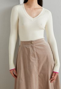 Vrouw in een crèmekleurige ribbel V-hals trui met roze biesjes op de mouwen en een hooggemiddelde beige ribfluwelen rok.
