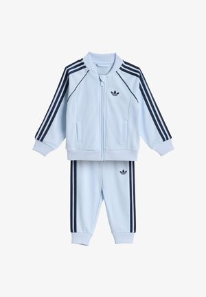 Chándal azul claro de dos piezas con franjas azul marino en las mangas y los pantalones, chaqueta con cremallera y logotipos de trébol de Adidas en el pecho y el muslo.