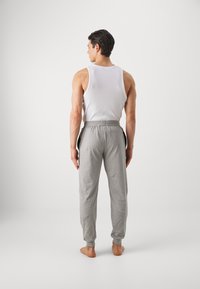 Pantalons de survêtement gris avec une taille élastique et des poches latérales, associés à un débardeur blanc. Le modèle est pieds nus et vu de dos.