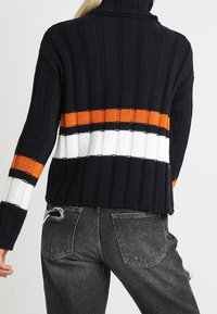 Stickad marinblå tröja med hög krage, prydd med horisontella ränder i orange, vitt och svart; ribbad textur, kort modell.