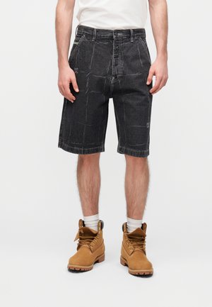 Mann trägt schwarze Jeansshorts, braune Arbeitsschuhe mit weißen Socken und ein weißes Hemd vor schlicht weißem Hintergrund.