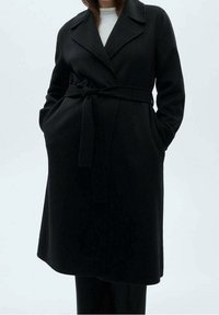 Manteau en laine noire avec taille ceinturée, grands revers, poches latérales et coupe décontractée. Texture lisse et design minimaliste.