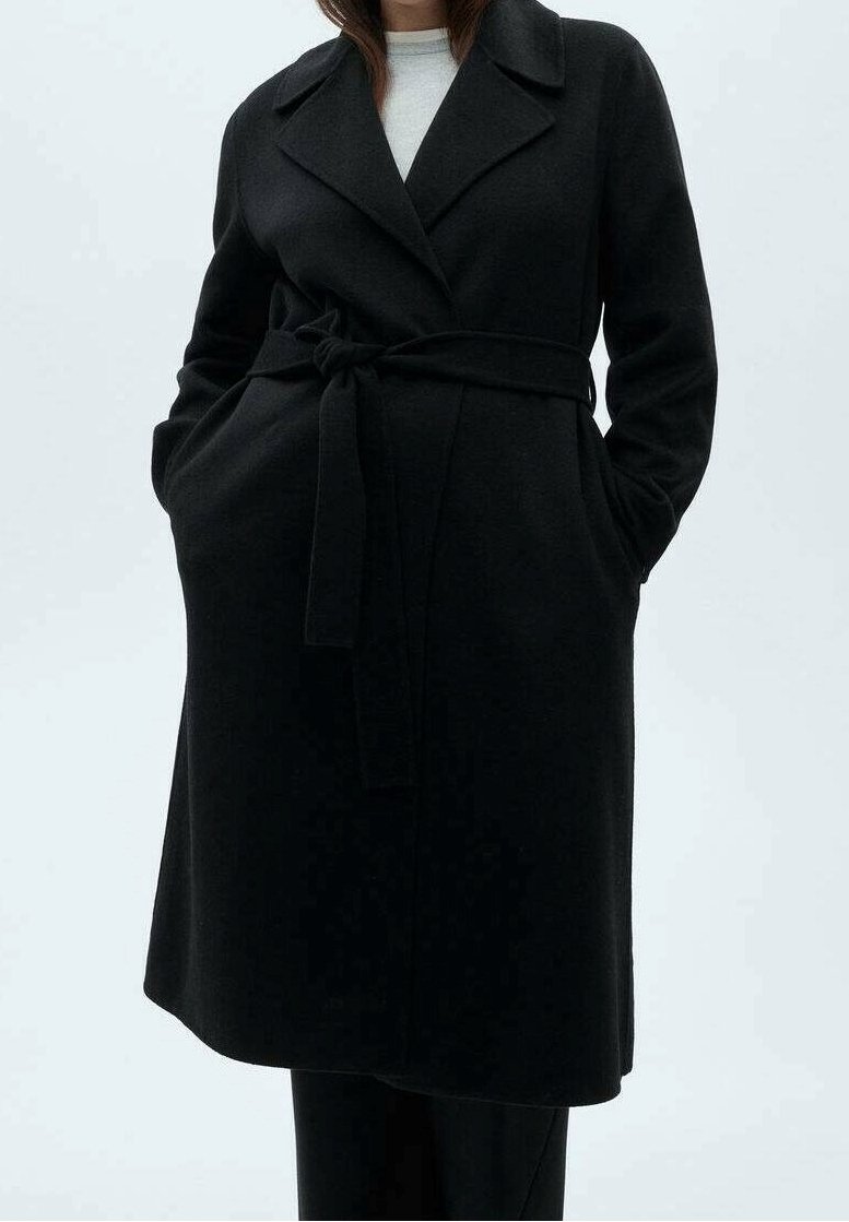 Manteau en laine noire avec taille ceinturée, grands revers, poches latérales et coupe décontractée. Texture lisse et design minimaliste.