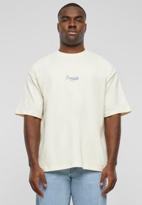 Cream-colored oversized cotton t-shirt with short sleeves, featuring a blue "Prohibited" graphic in cursive on the front.