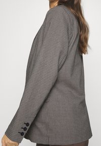 Veste à carreaux marron et noire avec une coupe ajustée, fermeture à un bouton, et trois boutons foncés sur le poignet ; texture de tissu lisse.