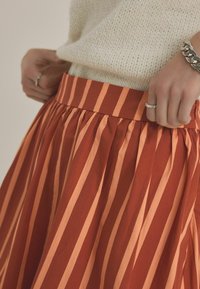 Een geplooide rok met verticale rode en lichtoranje strepen, voorzien van een effen tailleband en een textiel, lichtgewicht stof.
