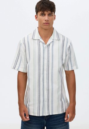 Joven con una camisa blanca de manga corta con rayas verticales grises y verdes, combinada con vaqueros azul oscuro.
