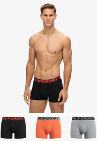 Bañadores ajustados negros con una banda elástica roja de "SUPERDRY". Tres pares adicionales en primer plano: naranja, gris y negro, todos con bandas similares.