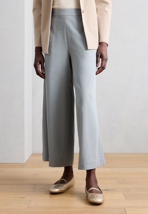 Osoba nosiaca svetlomodré široké nohavice, béžové sako a zlaté balerínky stojaca na drevenej podlahe pri obyčajnej stene.