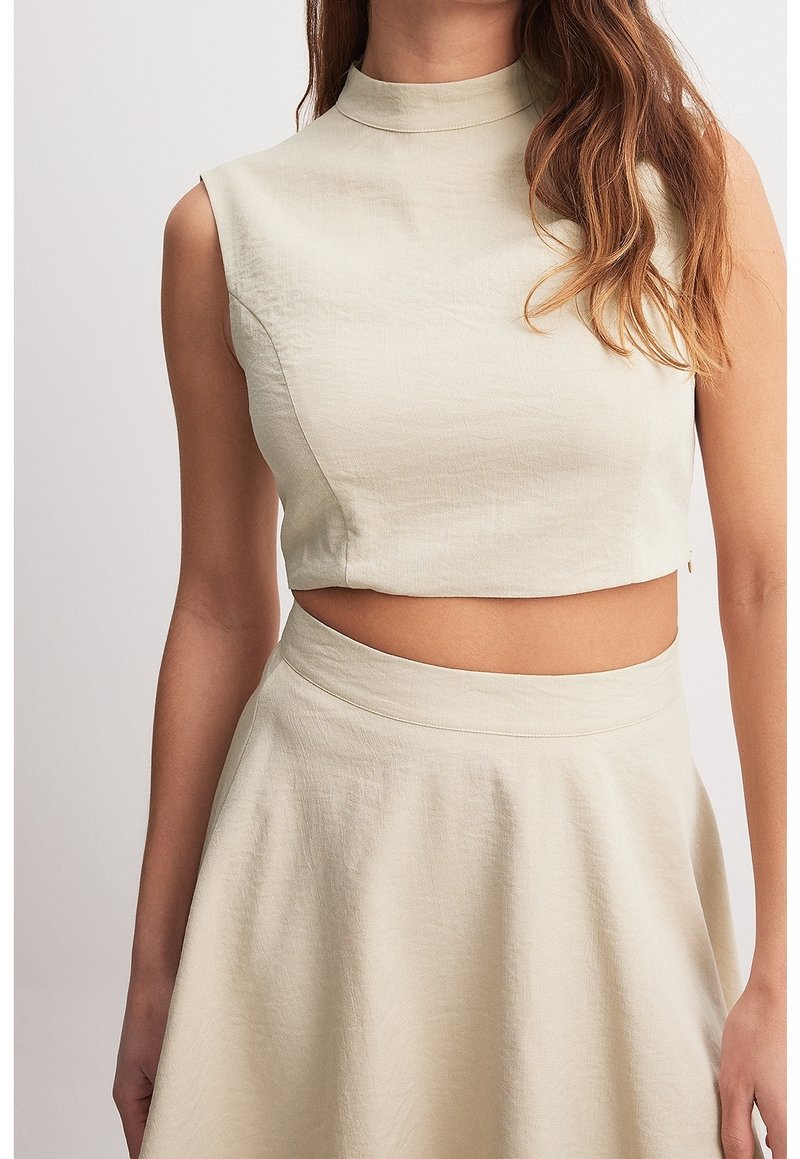 Beige cropped top met een hoge hals en mouwloos ontwerp, gecombineerd met een bijpassende hooggetailleerde rok met een gladde textuur.